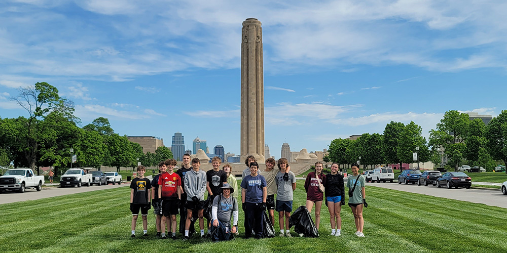 Modern photograph of a group of kids standing on the lawn with the Liberty Memorial Tower in the background, with large trash bags in hand